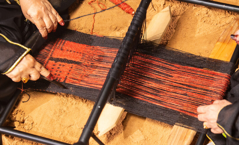light-skinned hands working on a loom strung with black and rust colored yarns
