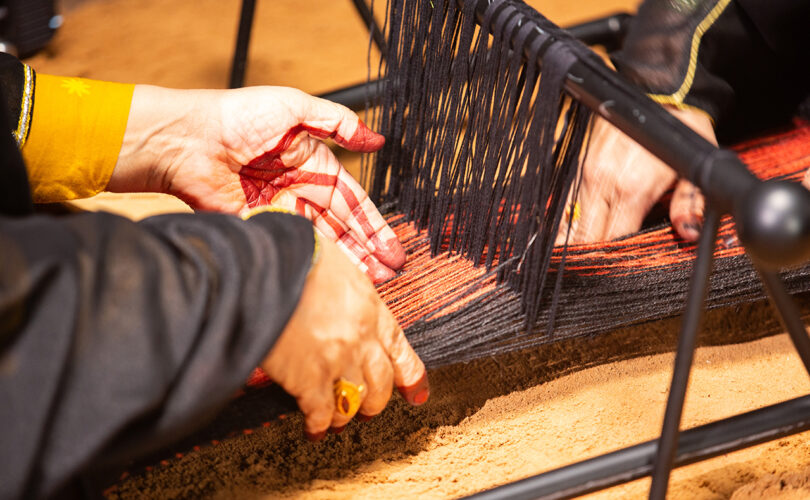 light-skinned hands working on a loom strung with black and rust colored yarns