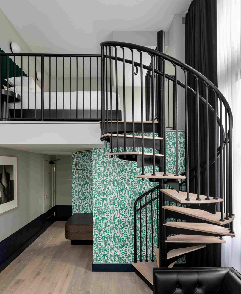 Loft spiral staircase leading to a bed. Walls are covered in green and white flora-themed wallpaper, with framed black and white photography on the left wall.