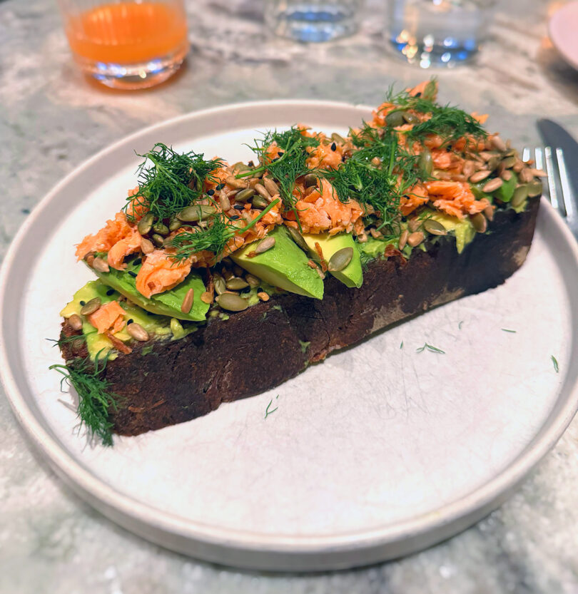 Large toasted slab of sourdough bread topped with slices of avocado, smoked salmon, pumpkin seeds, and garnished liberally with sprigs of dill.