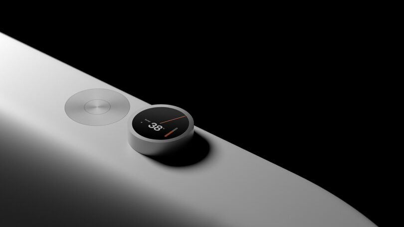Detail of the minimalist sculpted bath tub's touch dial control display, showing the temperature of the bath water. A metal push button sits flush along the tub surface on the left side of the dial.