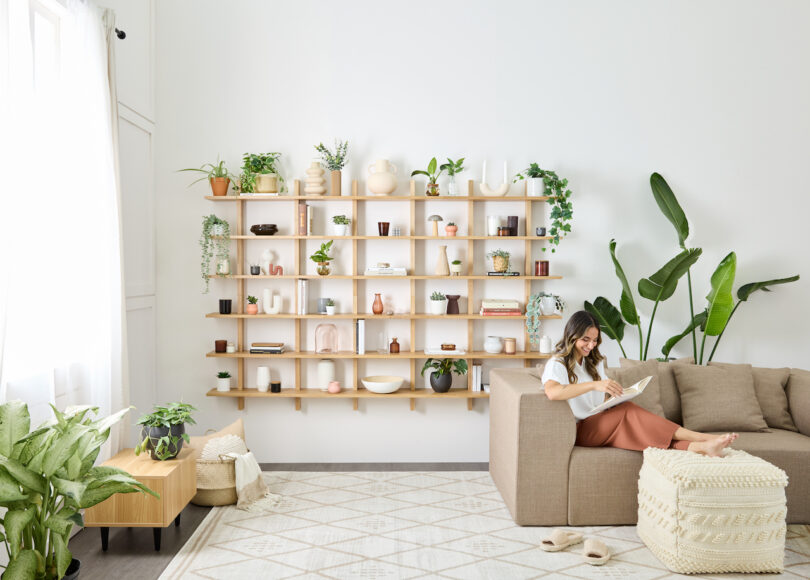 modular oak shelf holding various plants, books, and objects next to woman reading a book on a sofa