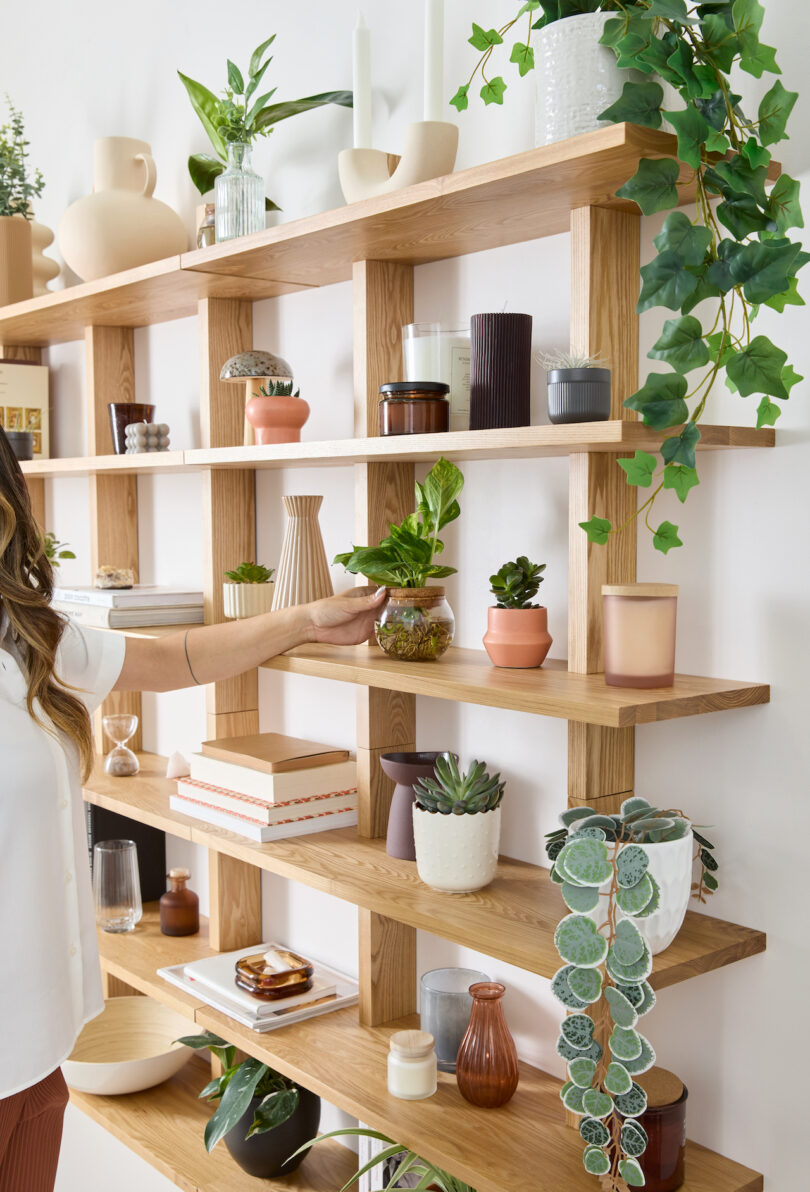 woman styling a modular oak shelf holding various plants, books, and objects