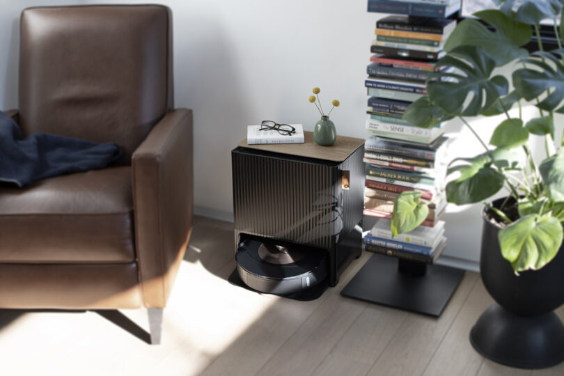 Roomba Combo j9+ robot vacuum and mop set in a living room setting in between a leather armchair and vertical book storage shelf to the right, alongside a large houseplant in a black planter.