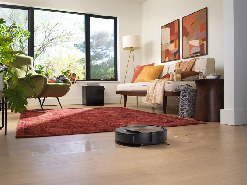 Roomba Combo j9+ robot vacuum and mop about to cross from hardwood floor over a red rug in a living room setting, with large three windows in the background and mid-century sofa to the right, house plants and arm chair to the left.