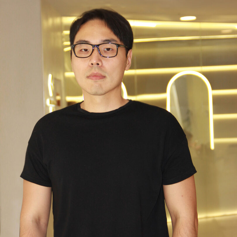 light-skinned man with dark hair wearing glasses and a black t-shirt looks at the camera