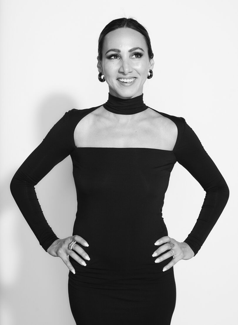 black and white portrait of a smiling woman with pulled back dark hair wearing a form-fitting black dress with her hands on her hips
