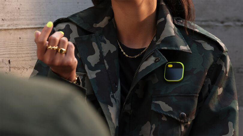 Woman wearing camouflage jacket and neon yellow nails wearing a human AI pin with a similarly colored yellow plastic casing around the device.