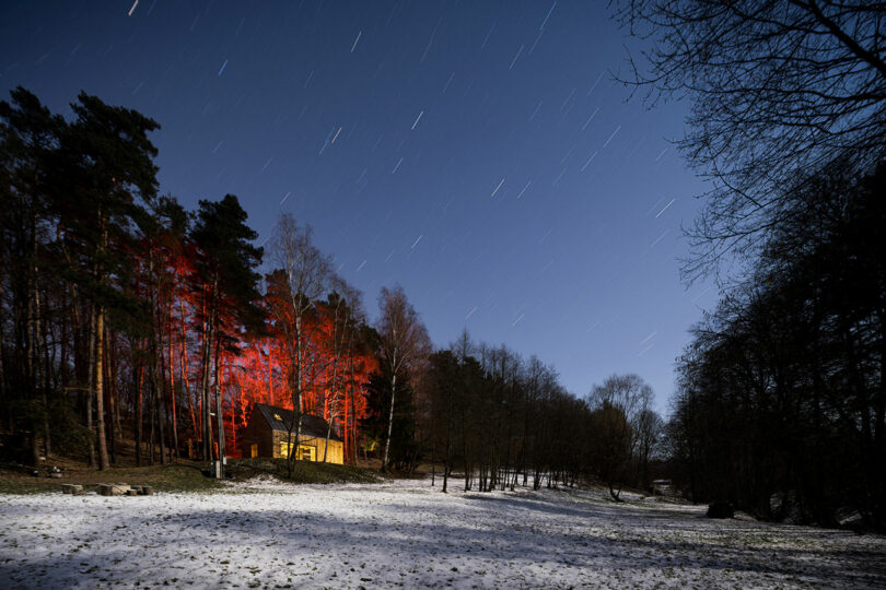 twilight angled view of modern wood cabin in woods with red lights shining on surrounding trees