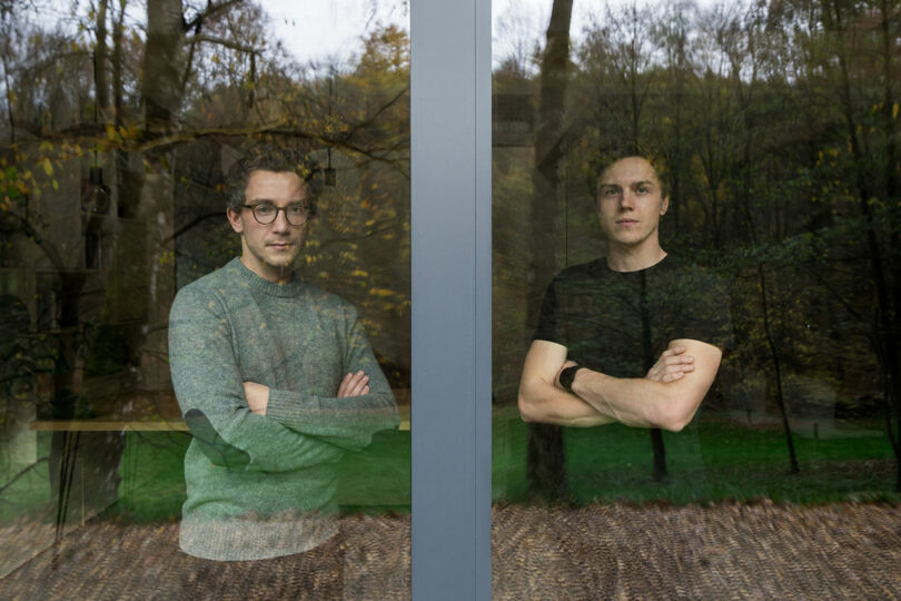 two men standing at angle towards each other with arms crossed looking at camera through window