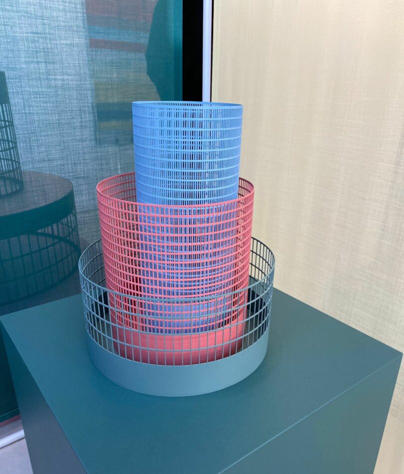 Three metal mesh cylindrical forms in teal, pink and blue, sit inside each other, each one narrower and taller than the one it sits inside. The collection sits on a teal plinth in front of a blue and a cream curtain at right angled to one another. 