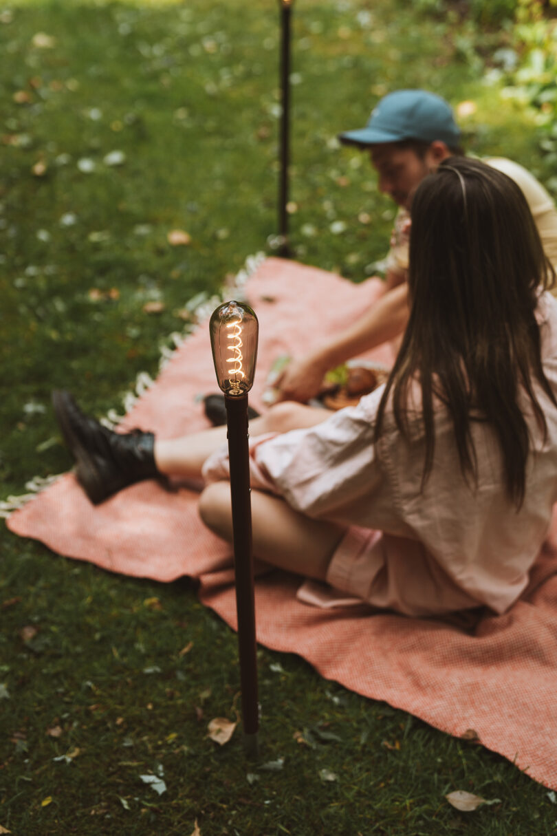 a couple sitting on a pink picnic blanket with two outdoor torches