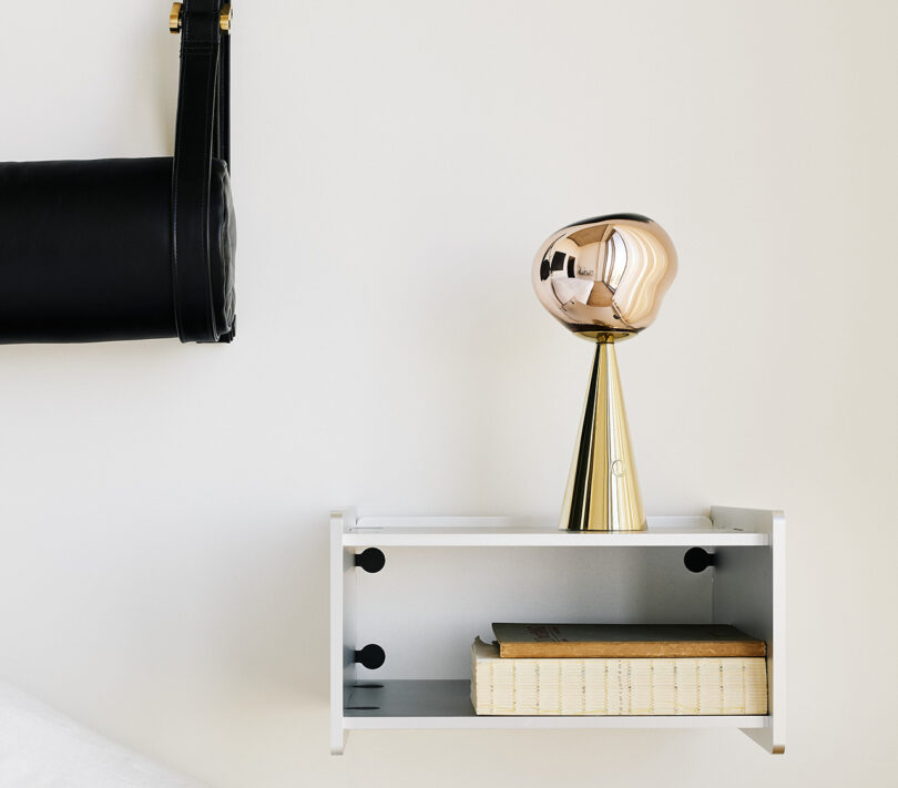 Side by side photos of Tom Dixon Melt portable rechargeable light in gold on a white floating bookshelf