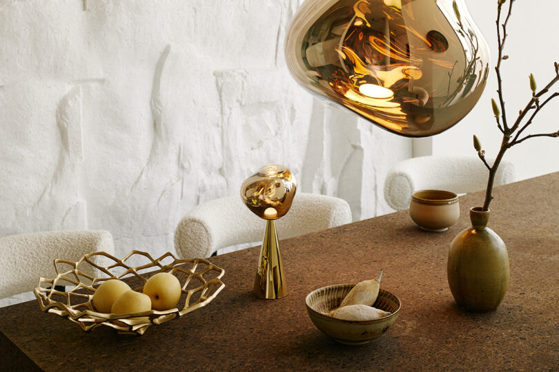 Side by side photos of Tom Dixon Melt portable rechargeable light in gold set upon a dining table alongside Melt pendant overhead and small tablescape accessories including a gold wire fruit bowl and three small ceramics.