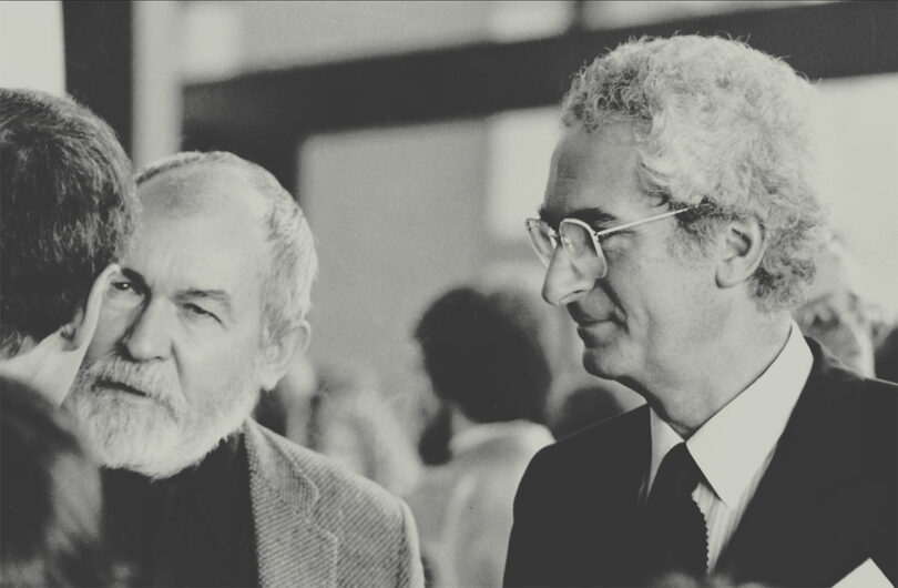 black and white photo of two older men with white hair talking to a man with back facing camera