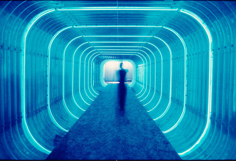 view into blue boxy tunnel with neon white lights wrapped around and a black figure in distance