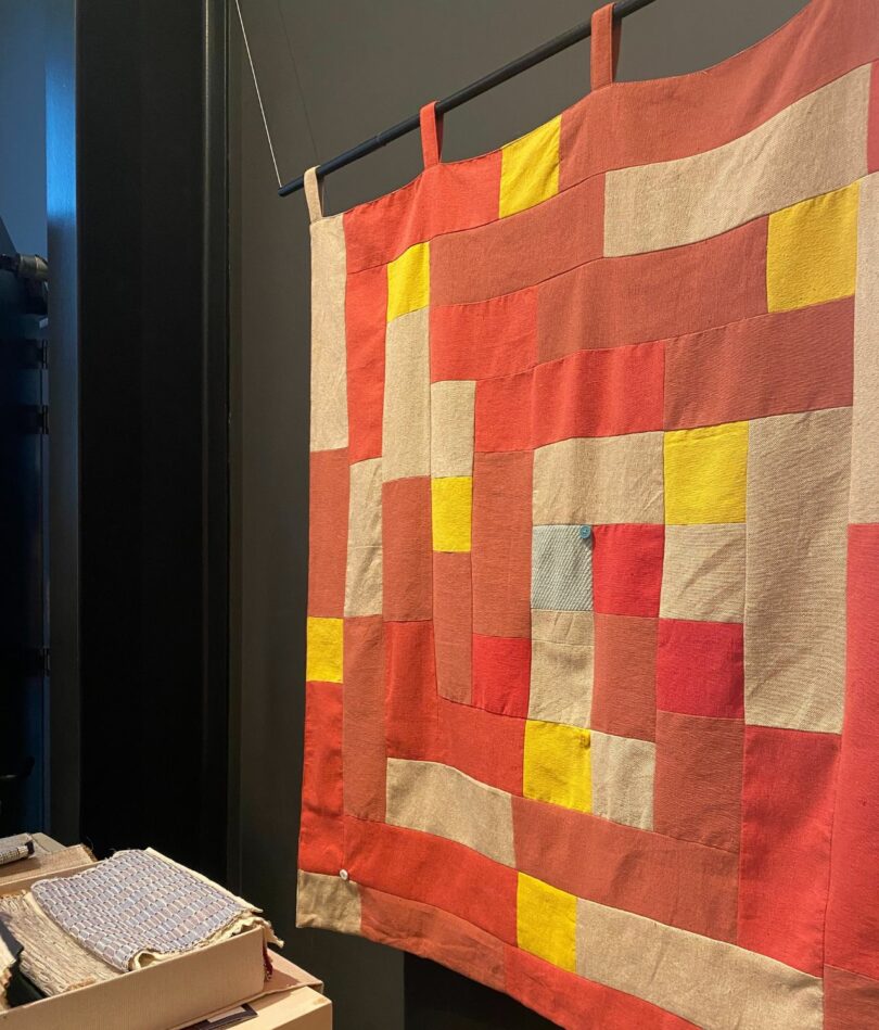 A wallhanging is made from patched together piece of fabric in various shades of red, orange, yellow and light brown with a pale blue square in the center. Buttons are sewn on at the point at which some of squares meet. 