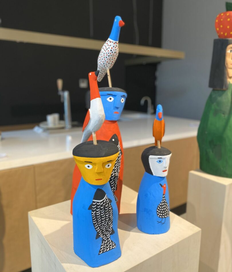 Wooden carvings of three brightly coloured, simplified, human forms each have a carved bird standing on their head and an illustration of either a bird or a fish on their fronts. 