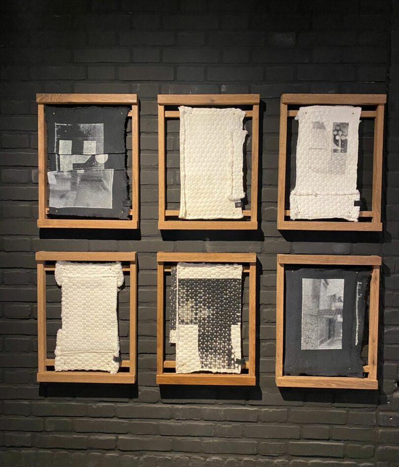 A series wooden frames are hung in two rows on a black wall. Black or white textiles that have been printed with black or black and white marks or images are stretched from the top to the bottom of each frame. 