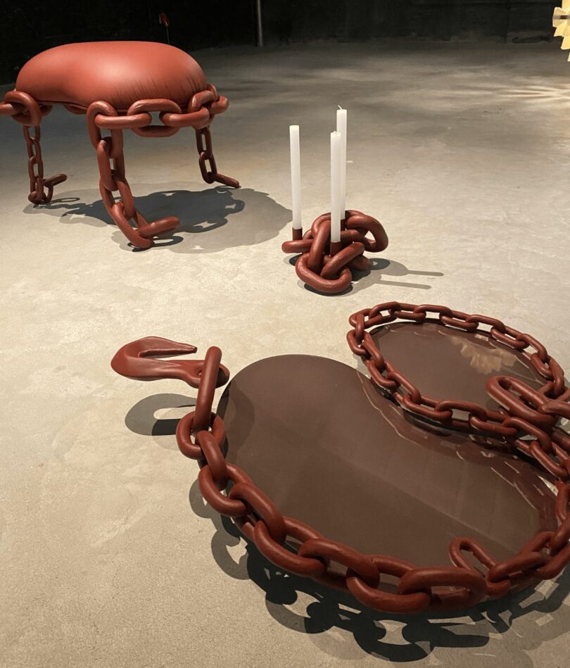 A stool, a candle holder and a mirror are on a concrete floor – they are all dark red and made primarily from oversized chains.