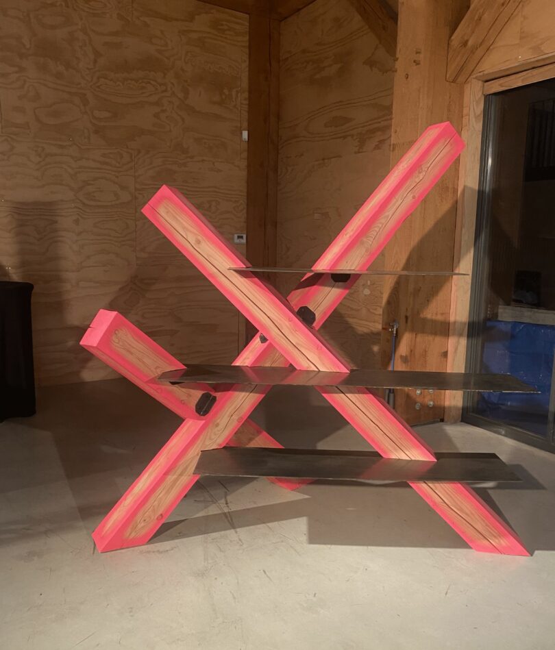 Three large wooden beams edged in neon pink are set at a 45 degree angle with the floor, so that they intersect each other. Horizontal polished metal shelves are inset into them at the front. 
