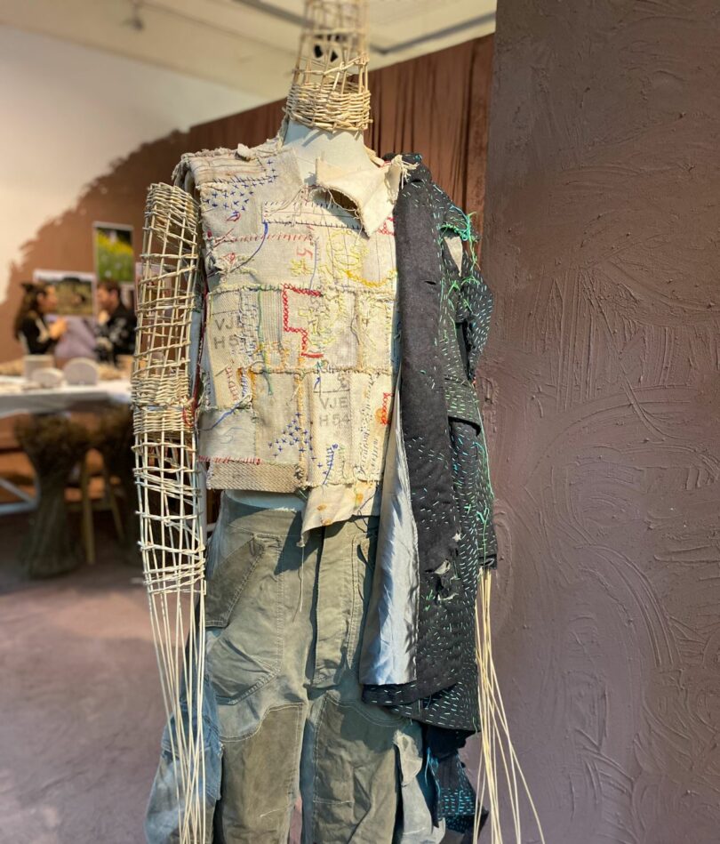 A mannequin with long pointy limbs and head is made from a straw mesh and wears pants, a sleeveless top and a jacket over one arm. The fabric of all three looks patched together and embroidered. 
