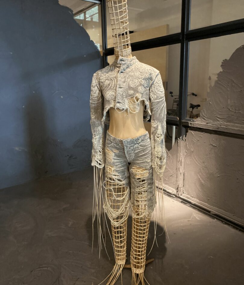 A mannequin with long pointy limbs and head is made from a straw mesh and wears a cropped jacket and torn trousers that look like denim embroidered with a floral pattern. 