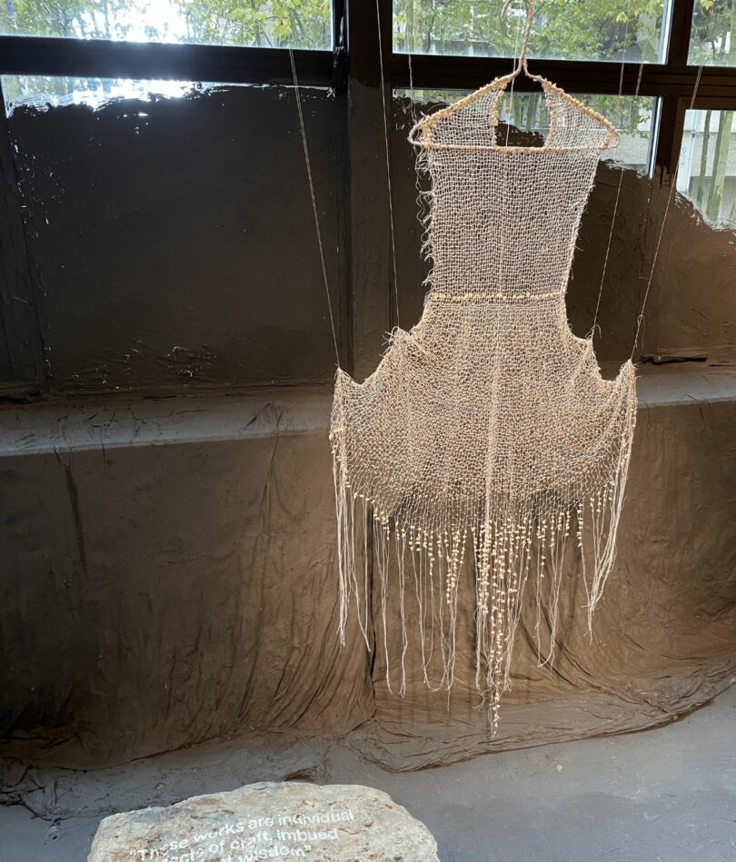 An incredible delicate woven dress is so fine as to be translucent. It hangs in front of a window, partly covered in clay.