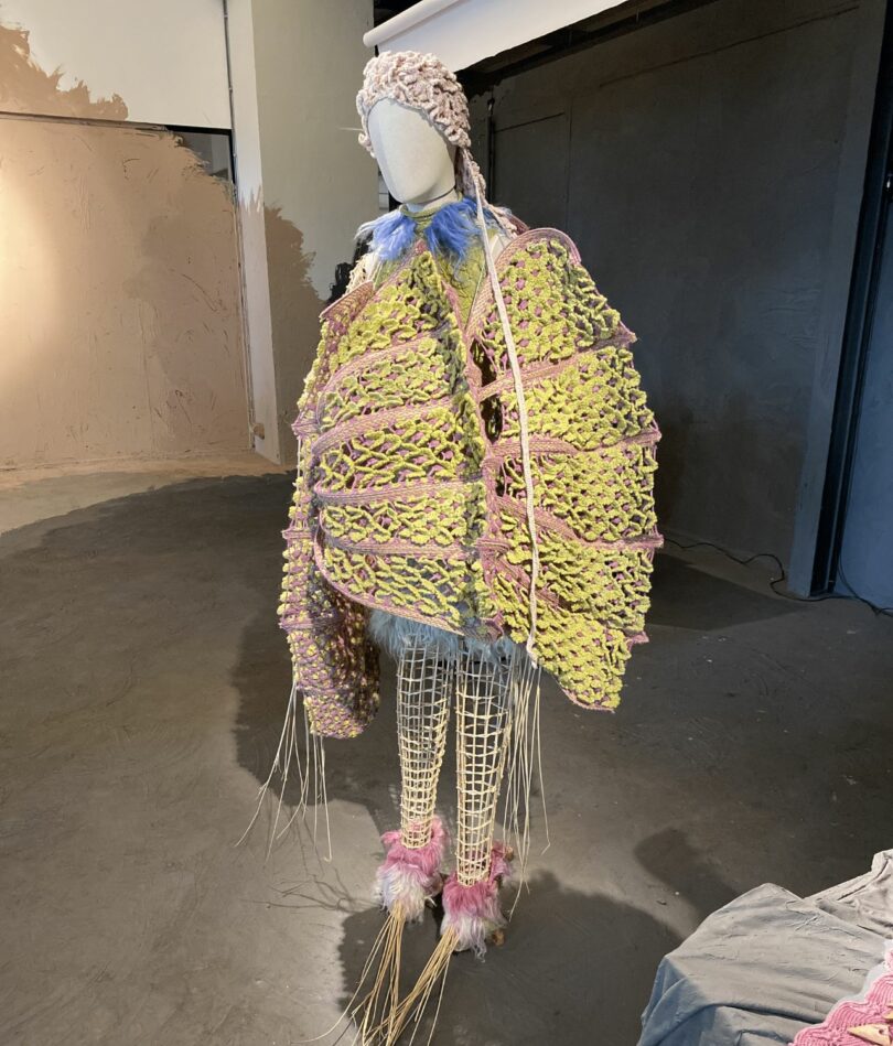 A mannequin with long pointy limbs has a typical faceless mannequin head and wears an oversized, structured garment in yellow and pink and what looks like a woven pink swimming cap. 