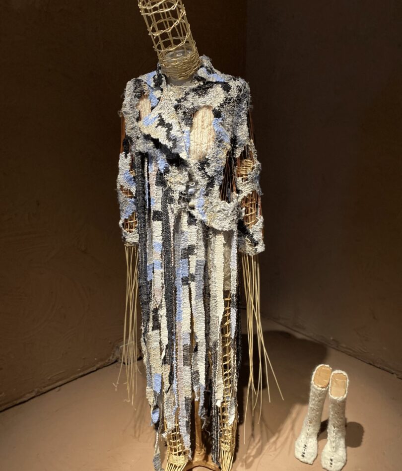 A mannaquin with long pointy limbs and head is made from a straw mesh and wears a long coat that looks tattered and is made from blue, white and black pieces. A pair of off-white woollen boots are on the ground to its left. 