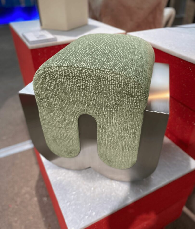 An ottoman has a "w" shaped metal base with a green "n" shaped upholstered foam section inserted into it. 
