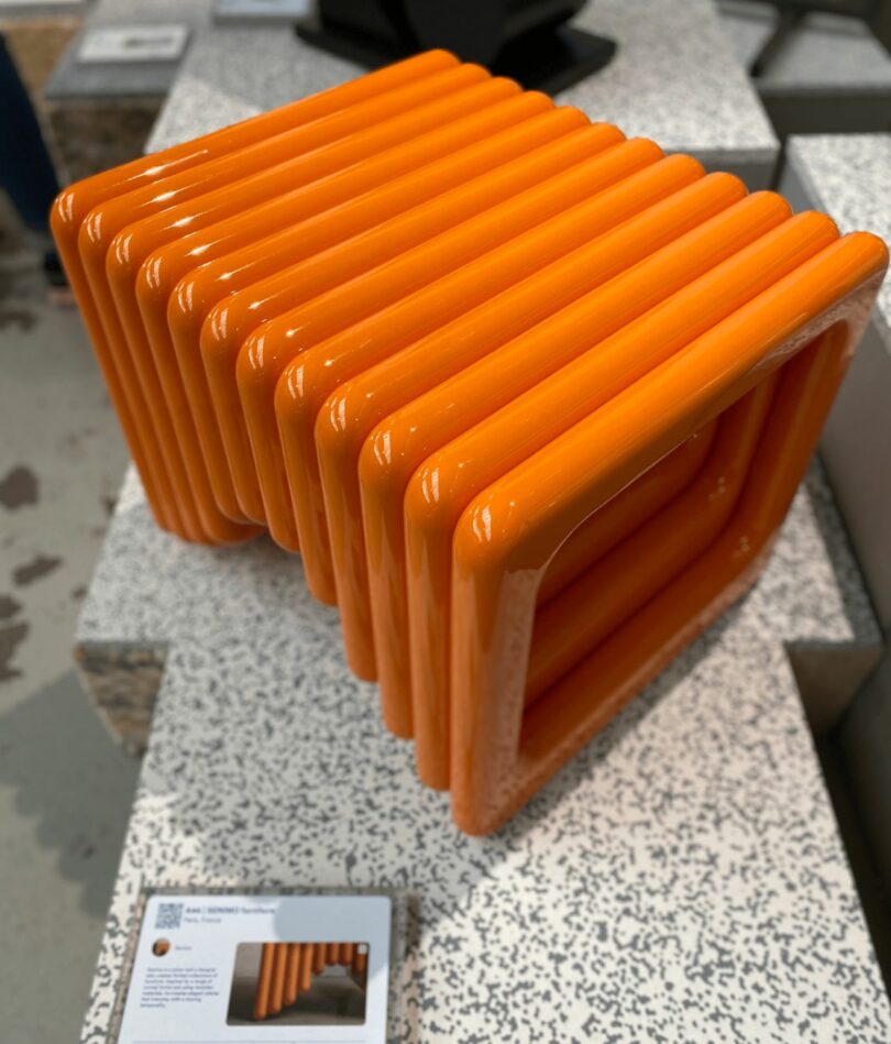 A side table or ottoman looks like it is made from a series of shiny, bright orange tubes arranged square loops