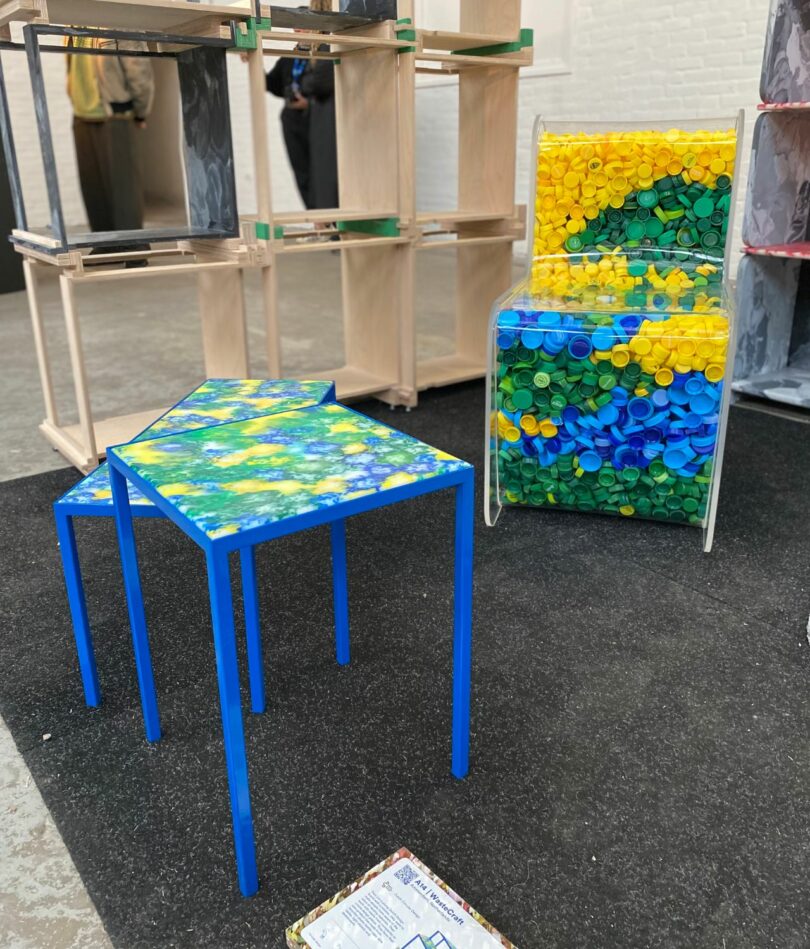 A clear plastic box filled with yellow, green and blue plastic bottle tops in behind two tables with bright blue legs and a surface made of swirled plastic in the same colour as the bottle tops