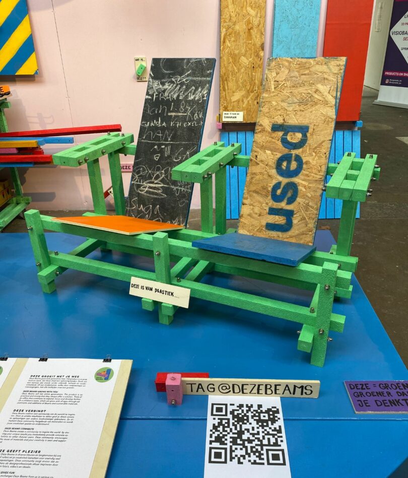 Two seats are made from a modular system of bright green beams and slats made from repurposed wood such as a chalkboard with white writing on it and an OSB board with the word "used" on it in blue
