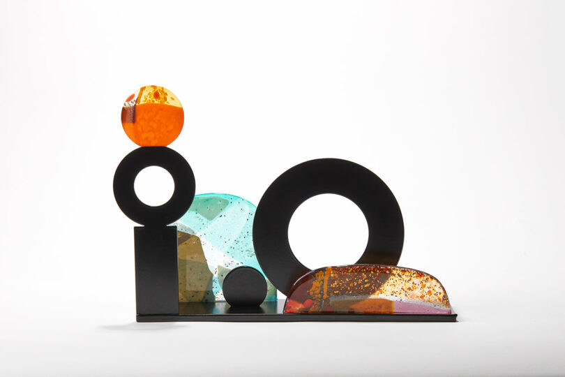A wide, mix media sculpture comprising black metal with orange and blue glass accents.