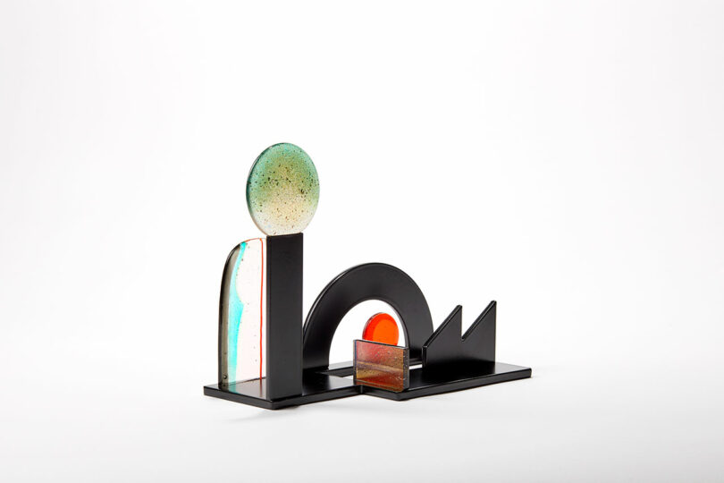 A wide, mix media sculpture comprising black metal with red and green glass accents.