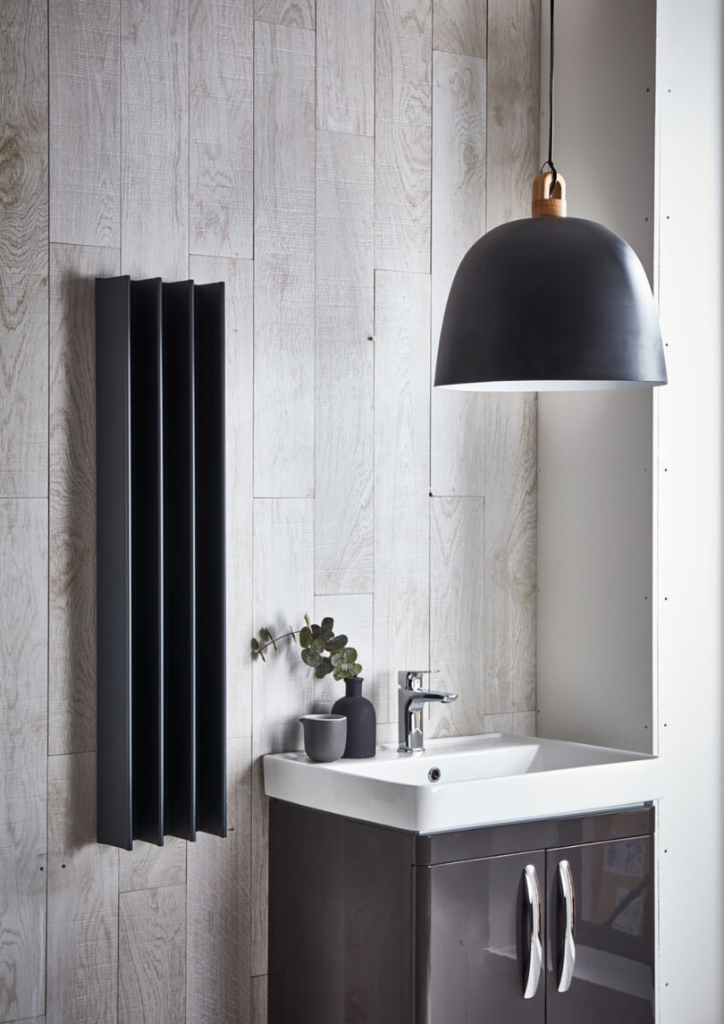 Modern bathroom with black vertical Gordon electric towel drying rail mounted on the left side near small sink basin with black pendant lamp overhead.