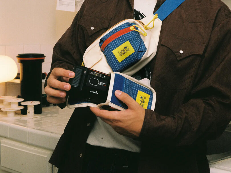 Man pulling an instant film camera from a Long Weekend camera carrying case, with another matching sling stretched diagonally across his torso.