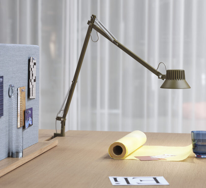 Olive green and brown pin mounted desk lamp with spring arm and small LED light illuminating a roll of yellow paper.