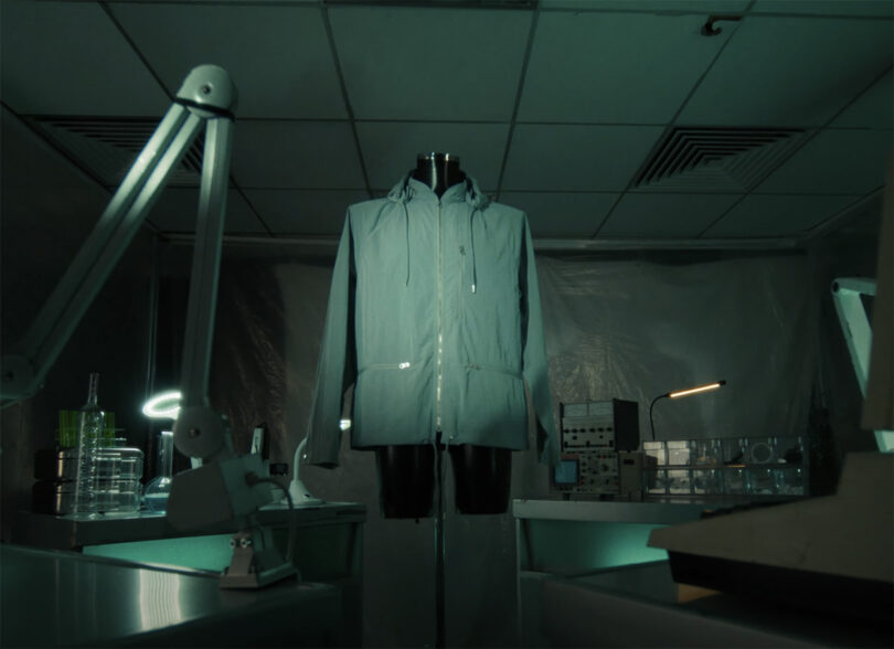 White semi-transparent labcoat designed by tech brand, Nothing, with zipper pockets and front, staged in a green cast laboratory setting.