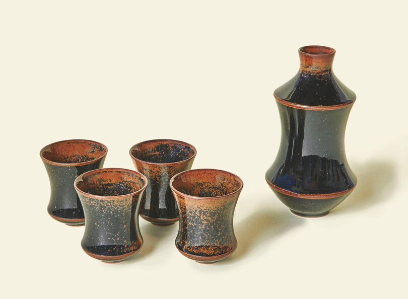 The Sake Set by Seth features a small ceramic carafe and matching cups with dark speckled and glossy glaze.