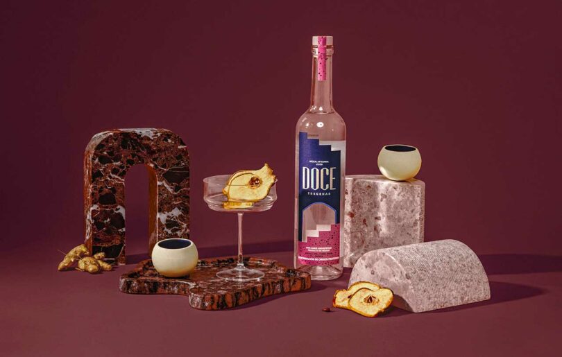 Doce Mezcal bottle and drinking glass staged against burgundy background with abstract ceramic shapes