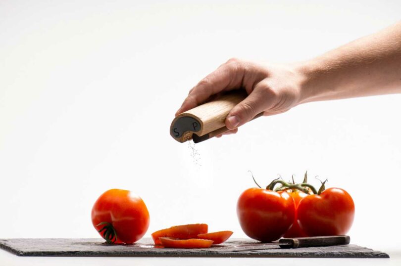 Person's hand holding minimalist salt and pepper shaker over sliced tomatoes