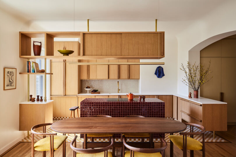 آشپزخانه مدرن گرم با جزیره آشپزخانه با کاشی بورگوندی تیره و قفسه بالای سر چوبی.  میز ناهارخوری با 6 صندلی میانه قرن در پیش زمینه.