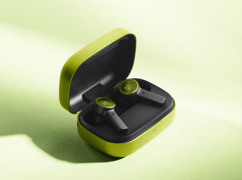 Beoplay EX wireless earbuds with an open bright lime green exterior case with black interior staged at an angle across a white surface illuminated with yellow-green lighting.