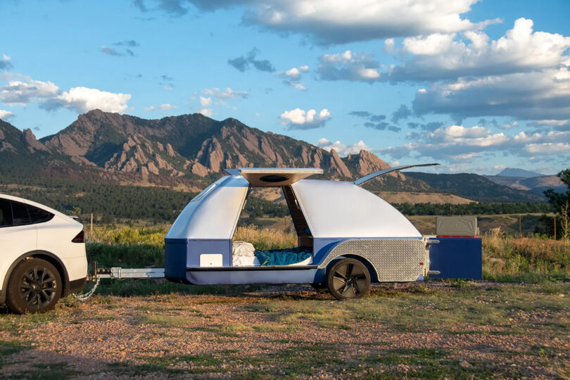 Silver and blue The Boulder teardrop trailer parked against the backdrop of rocky arid mountains and partially cloud skies.