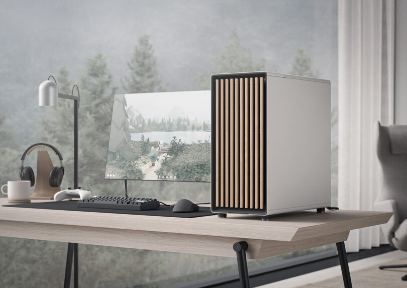 Fractal gaming PC with white case and slat wooden front on desk with monitor, headphones, lamp, wireless gaming control pad and mouse nearby.