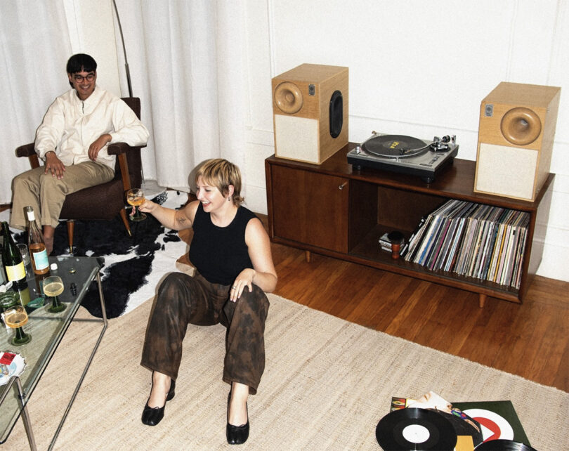 Young man with bowl cut in armchair and young woman with short blond hair laughing holding cocktail seated on ground listening to music from a turntable and two bookshelf speakers.