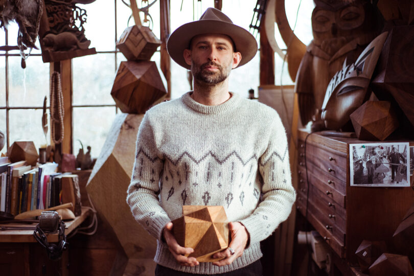 Sculptor Aleph Geddis in his carving shed holding up a polygonal shaped wood carving.