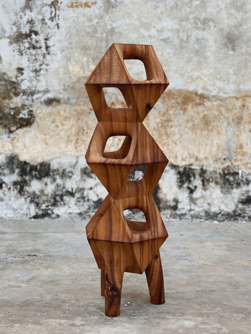 Vertical wood carved totem sculpture by Aleph Geddis, standing upon 3 leg base.